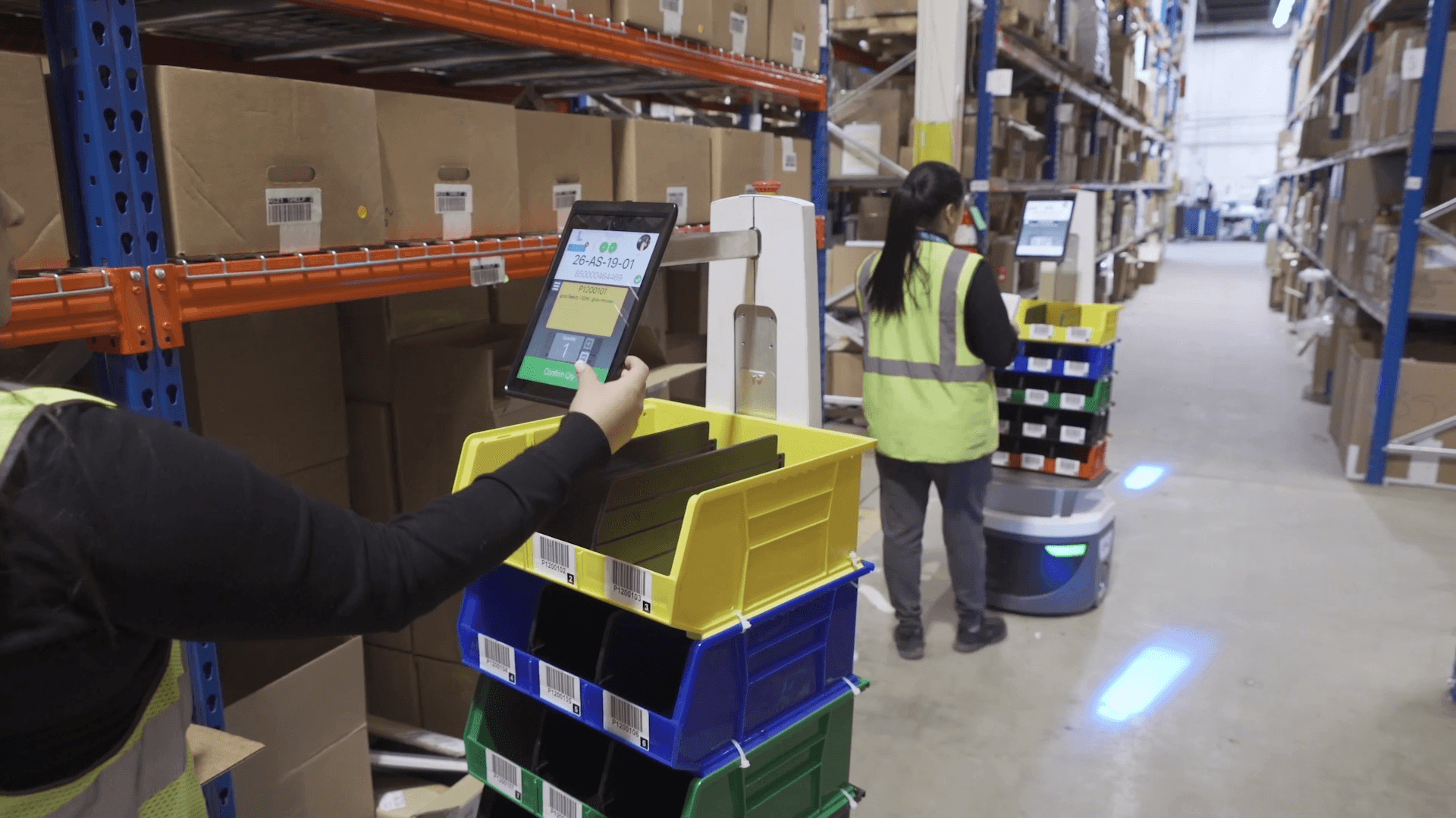 Warehouse workers, wearing safety vests, interact with robotic carts carrying colorful bins. They are surrounded by shelves stacked with cardboard boxes, creating an organized industrial setting.