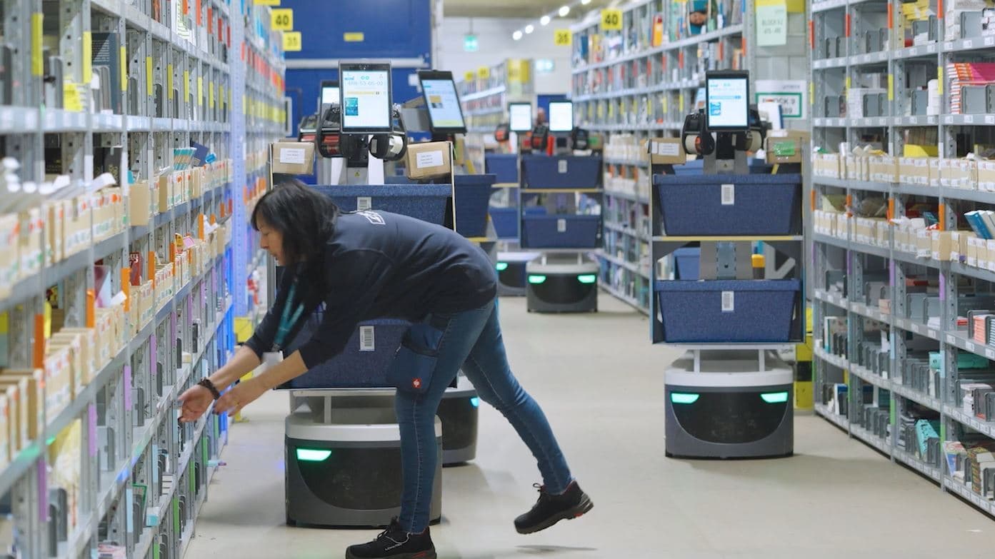 A person is interacting with shelves in a large warehouse, assisted by three robotic carts. The aisles are filled with various products, and electronic screens are visible on the carts, indicating a busy and automated environment.