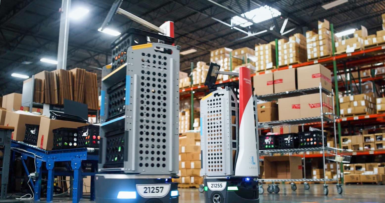 Two automated robots navigate an industrial warehouse, carrying stacked boxes. The background shows shelves filled with cardboard packages, and a conveyor belt on the left. The setting is well-lit with a high ceiling and visible structural beams.