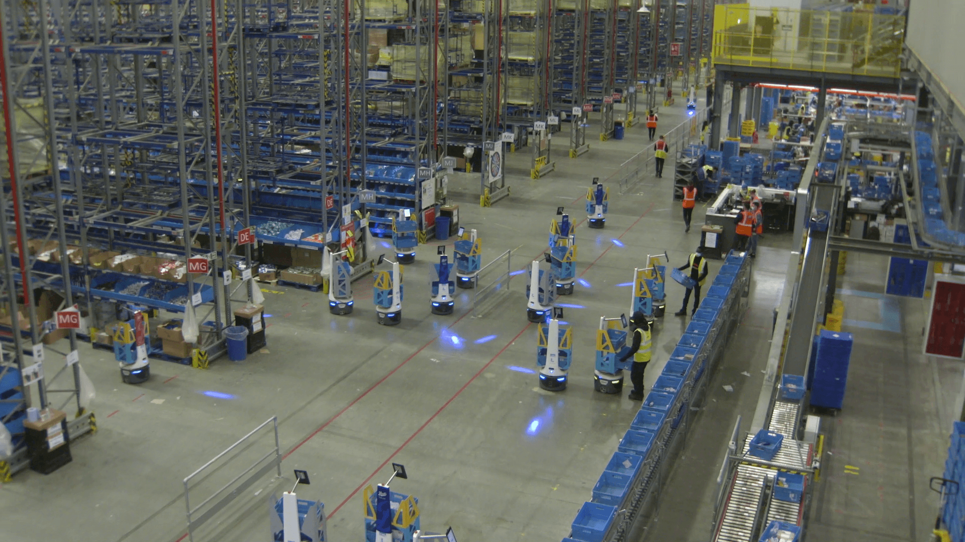A large warehouse with automated robots moving along the floor, surrounded by tall shelves filled with goods. Workers wearing safety vests are visible, overseeing operations. The environment is organized and busy, with a focus on efficiency.