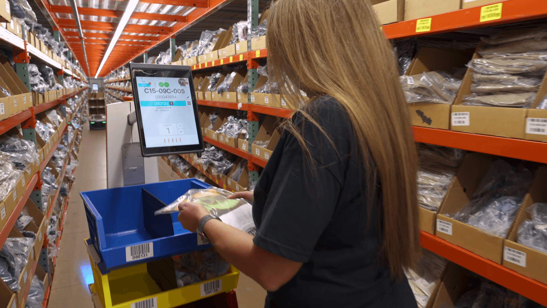 Person with long hair scans items at a computer station in a warehouse aisle filled with shelves of packaged goods. A blue bin is on a cart beside them, and the shelves are labeled with numbers and letters.