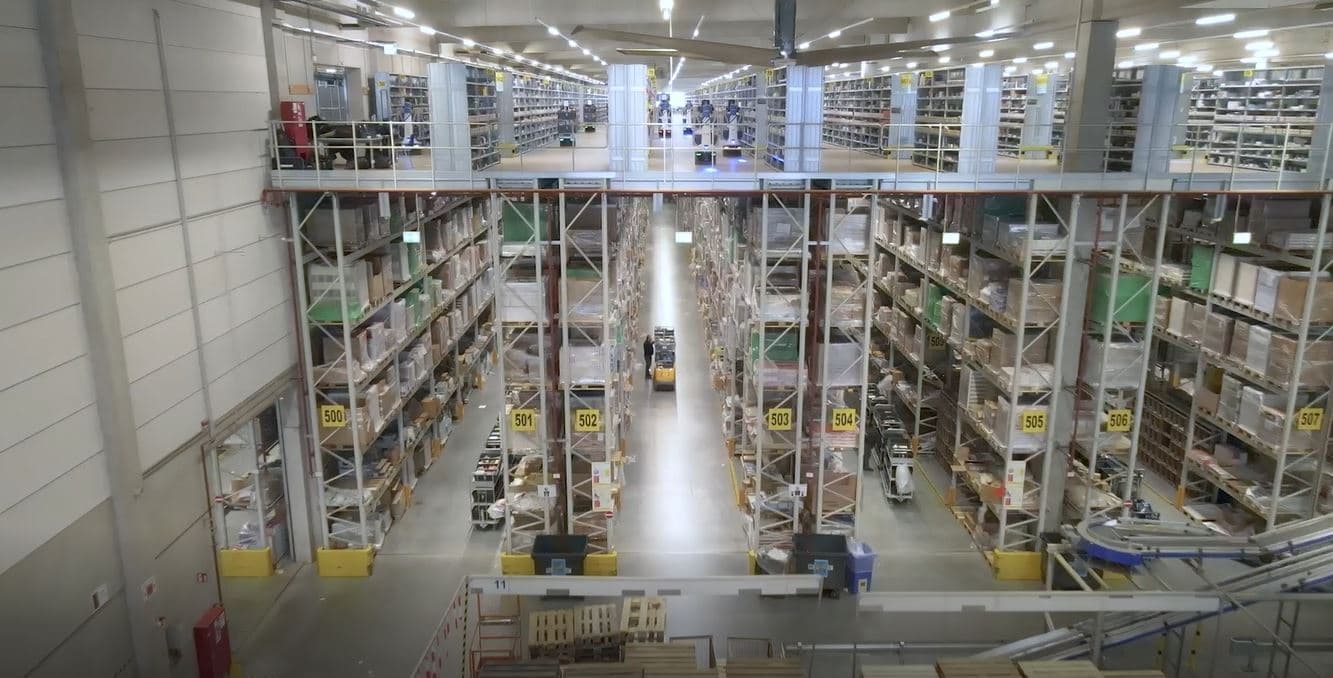 Wide-angle view of a large warehouse interior with tall shelves stacked with various boxes and items. A forklift is visible in the central aisle. Overhead lights illuminate the spacious, organized storage area.