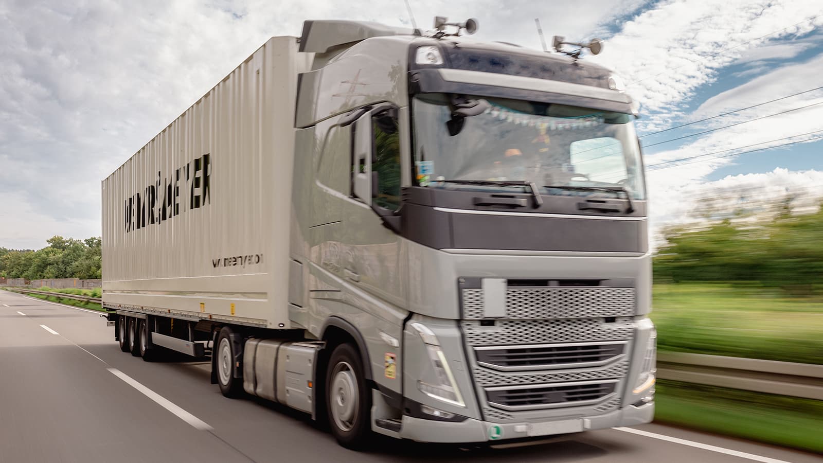 A large, modern semi-truck with a white trailer drives along a highway with a blurred background of greenery and a blue sky with clouds. The truck features high-tech equipment on its roof.