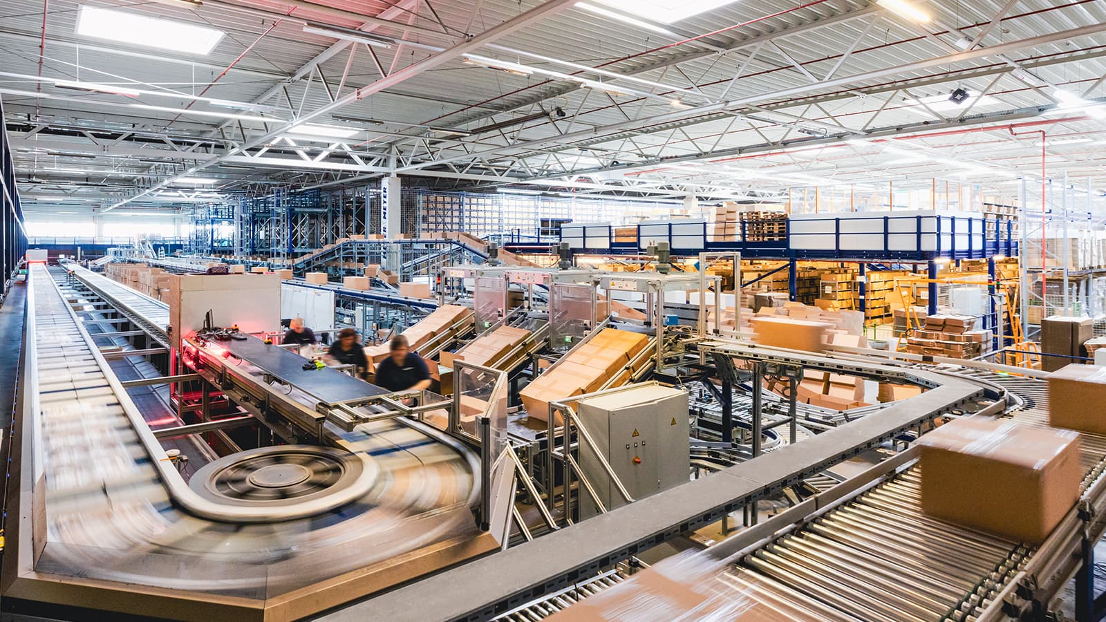 A large warehouse with automated conveyor belts transporting boxes and packages. Workers are visible moving among the machinery. The space is well-lit, with shelves of inventory in the background and a high ceiling with metal beams.