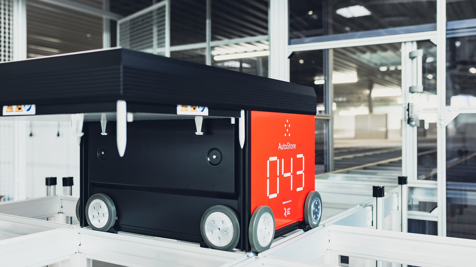 A red and black warehouse robot with wheels is moving on a track system within an industrial setting. The robots display shows the number 043. The environment is well-lit with glass panels in the background.