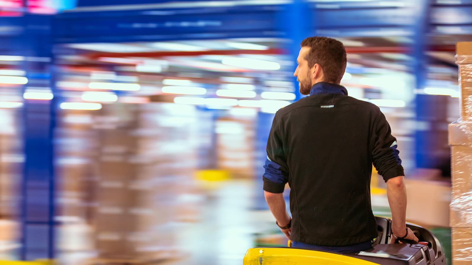 A person wearing a black sweatshirt operates a yellow pallet jack in a busy warehouse with motion blur. The background shows blue structures and stacks of boxes, indicating a fast-paced logistics environment.