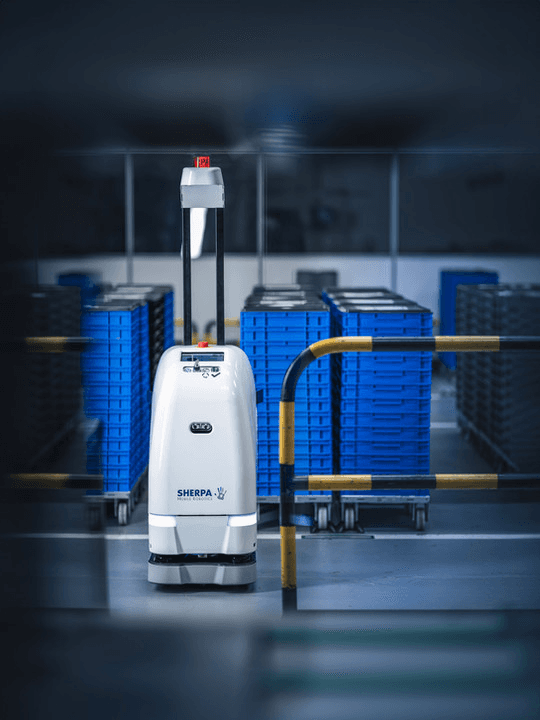 A white robot with the label SHERPA is stationed in an industrial setting. It stands on a gray floor, surrounded by stacks of blue containers on wheels. A yellow and black safety barrier is in the foreground.
