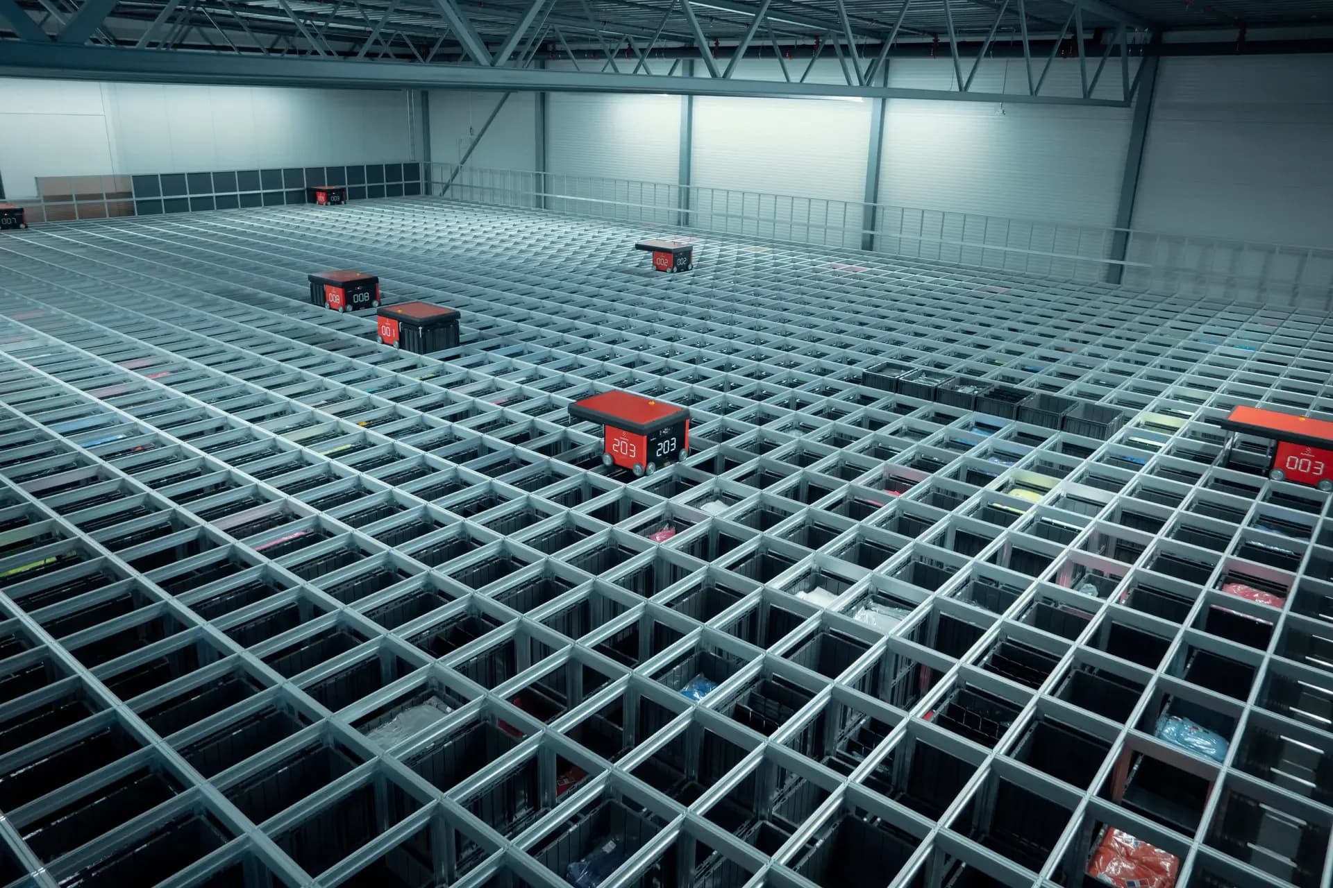 A large automated warehouse system with rows of metal grids and several red robotic bins on top. The structure is well-lit, showcasing the industrial setting and the organized storage design.