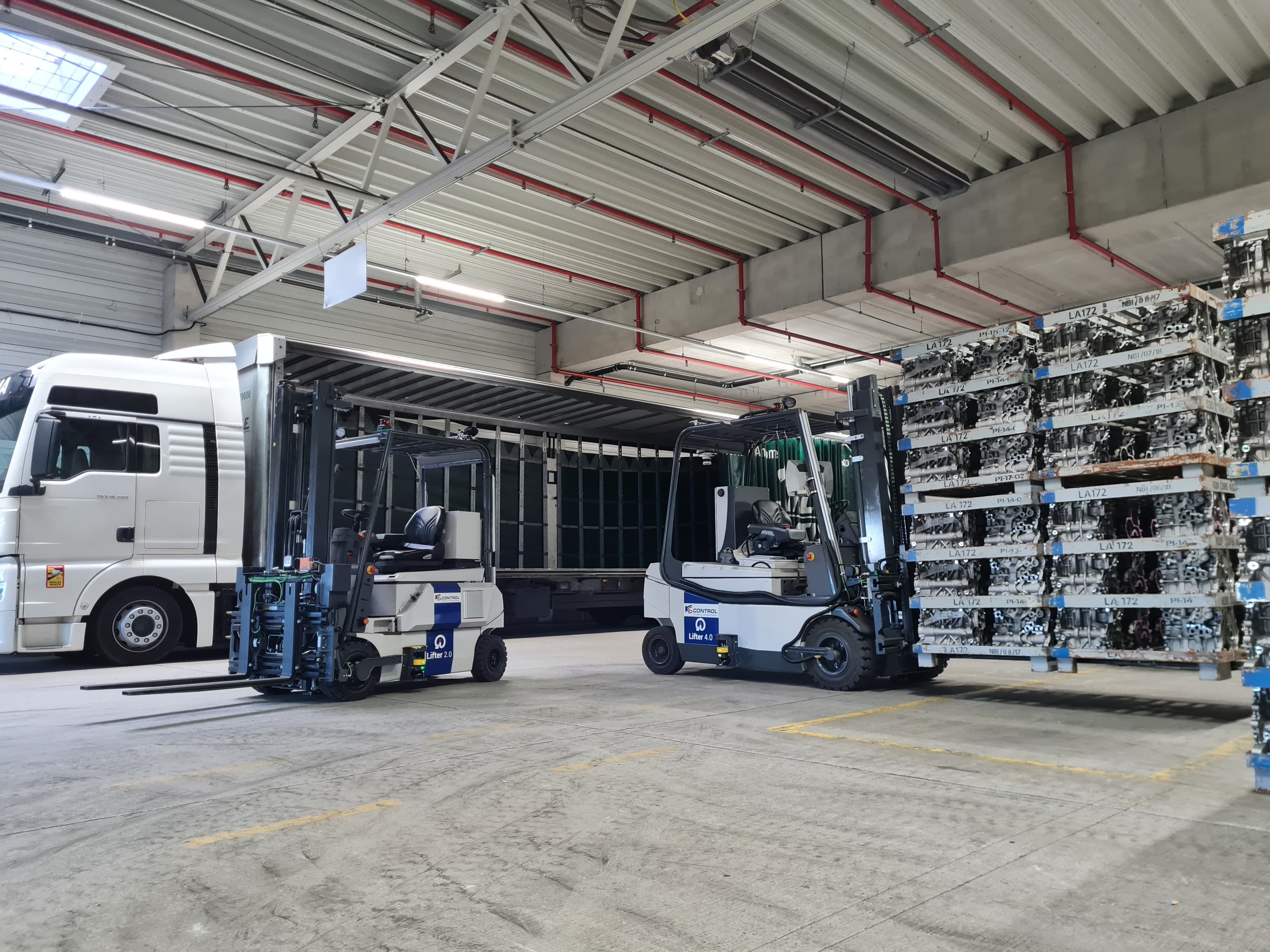 Two forklifts in a warehouse unloading metal racks from a white truck. The space has a high ceiling with visible beams and overhead lighting. The scene is organized and industrial.
