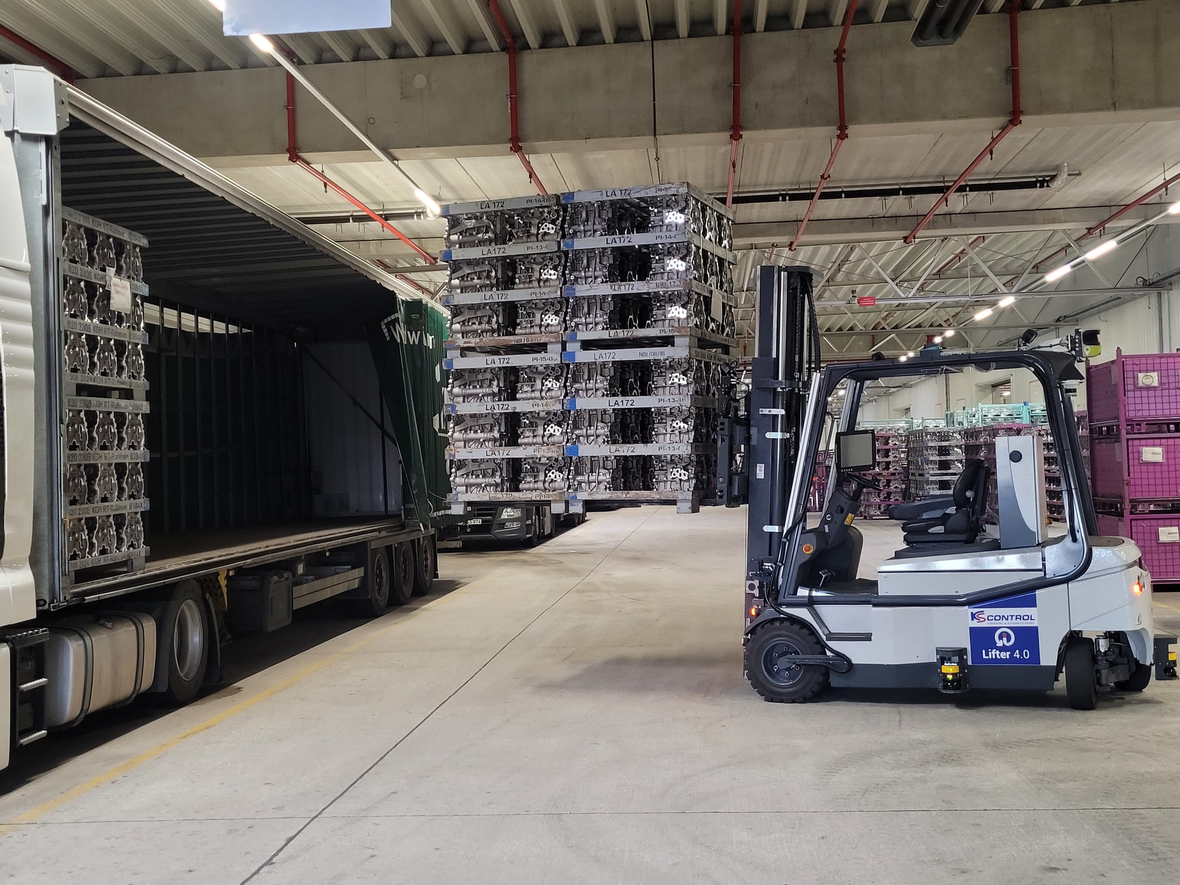 A forklift is parked inside a large warehouse next to a trailer being loaded with stacked metal crates. The warehouse has a high ceiling, visible pipes, and a concrete floor. Large stacks of crates are in the background.