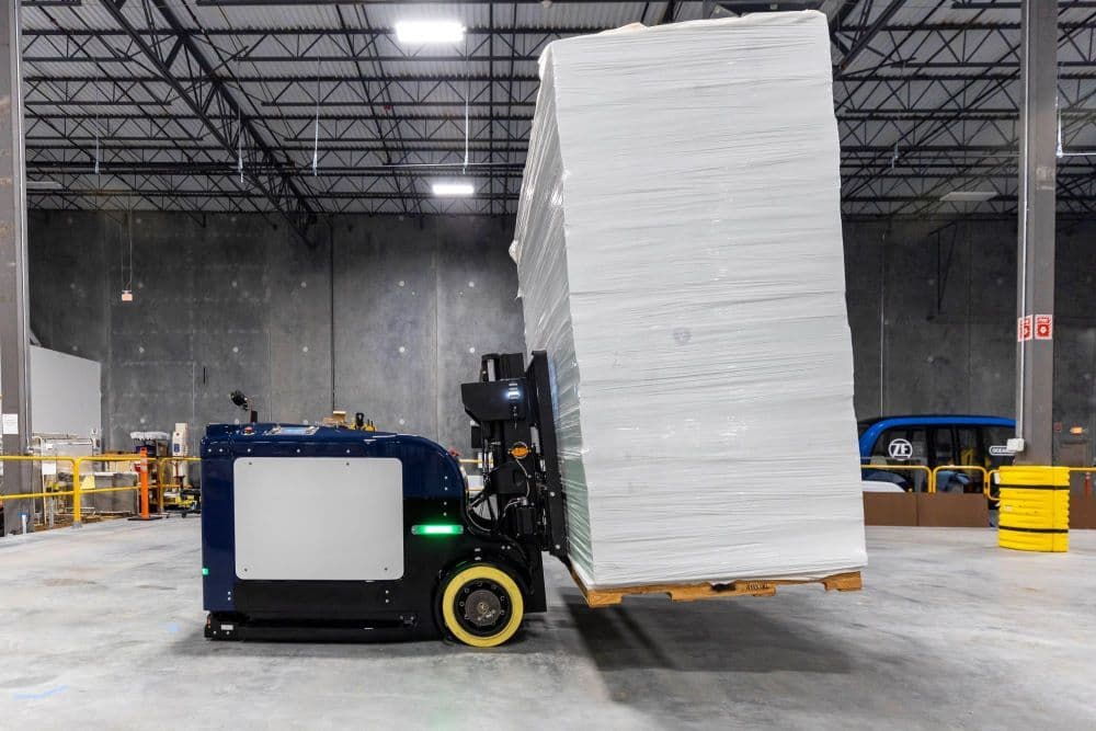A blue autonomous forklift transports a tall stack of white, wrapped goods on a wooden pallet inside a large warehouse with high ceilings and industrial shelving.