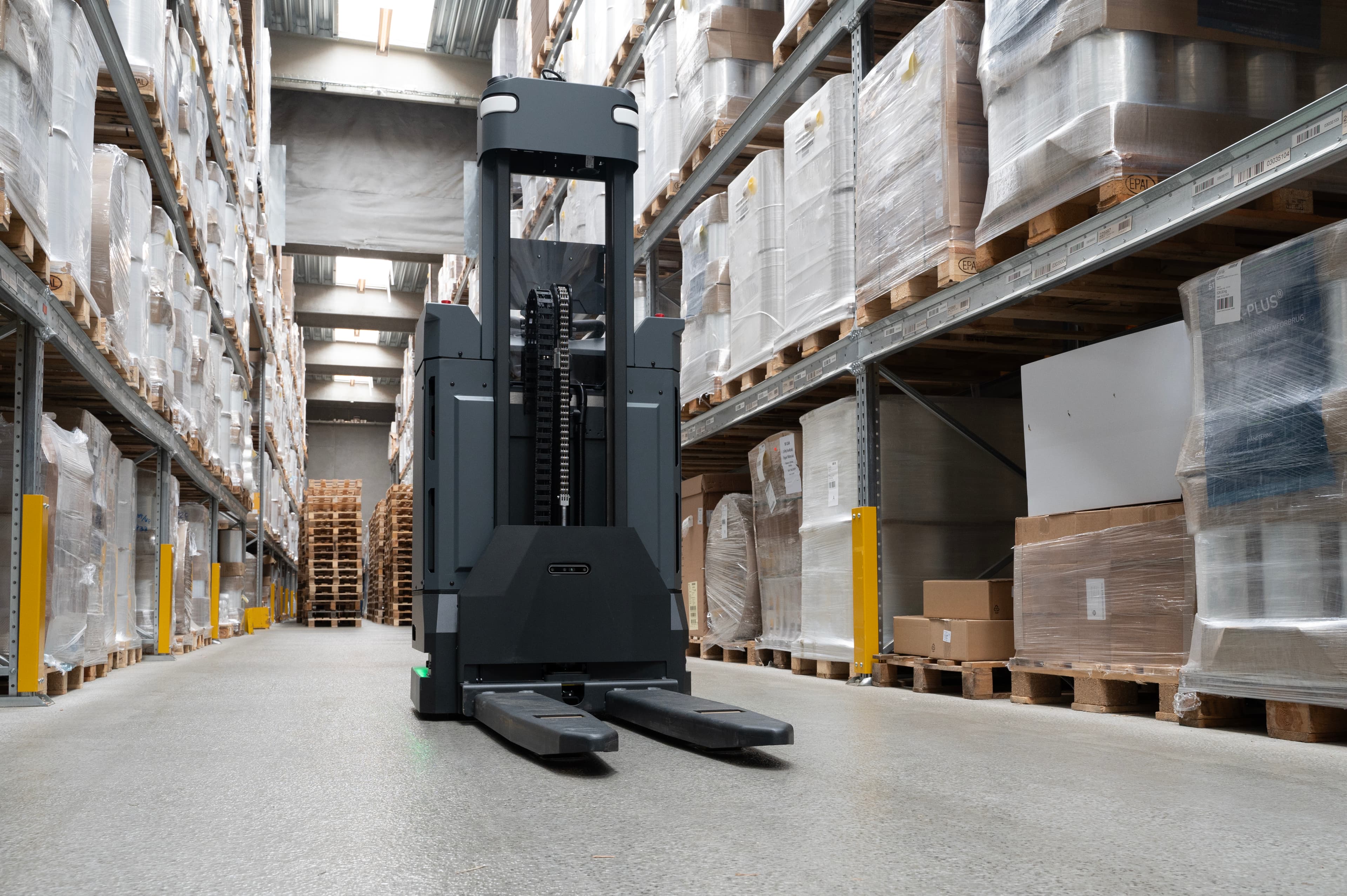 A modern forklift is parked in the aisle of a large warehouse, surrounded by shelves stocked with stacked, wrapped pallets and assorted boxes. The ceiling is high with natural light streaming in.