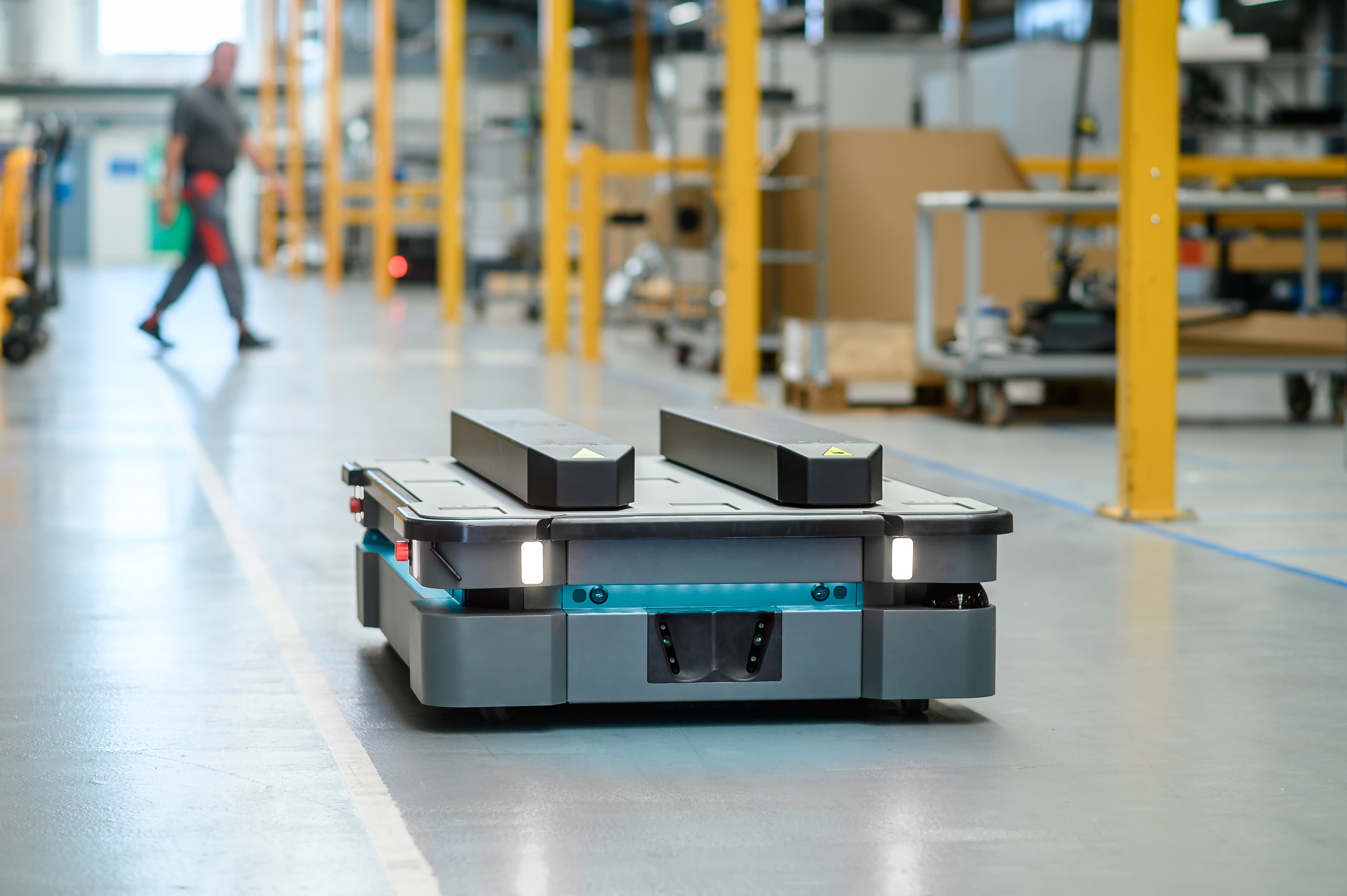 A robotic platform with sensors navigates a clean, spacious warehouse environment. Yellow support beams and industrial equipment are visible in the background, with a blurred person walking in the distance.