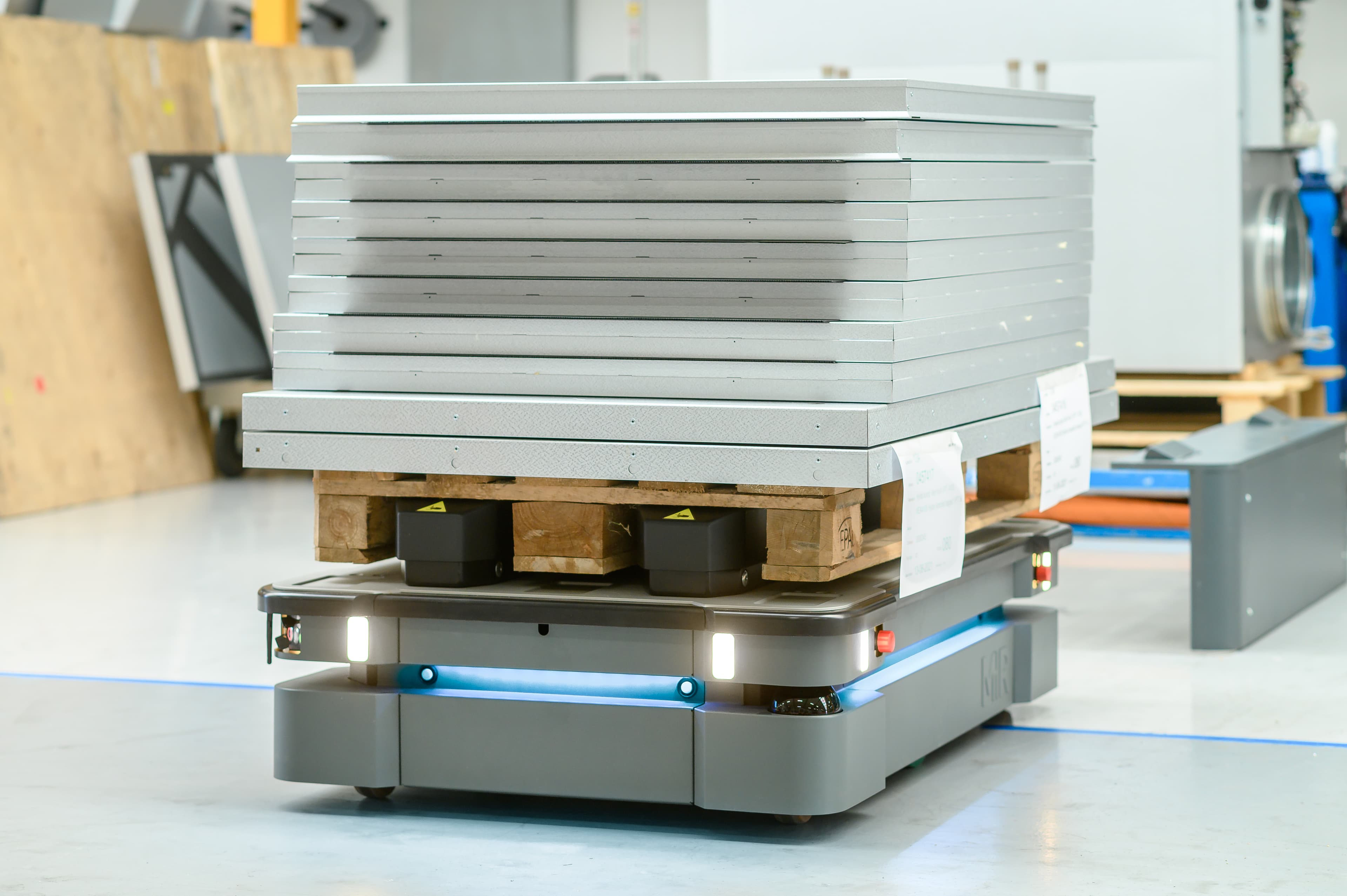 A robotic platform transports a stack of metal sheets on a wooden pallet across an industrial floor. The robot features LED lights and sleek, modern design elements suitable for automated logistics.