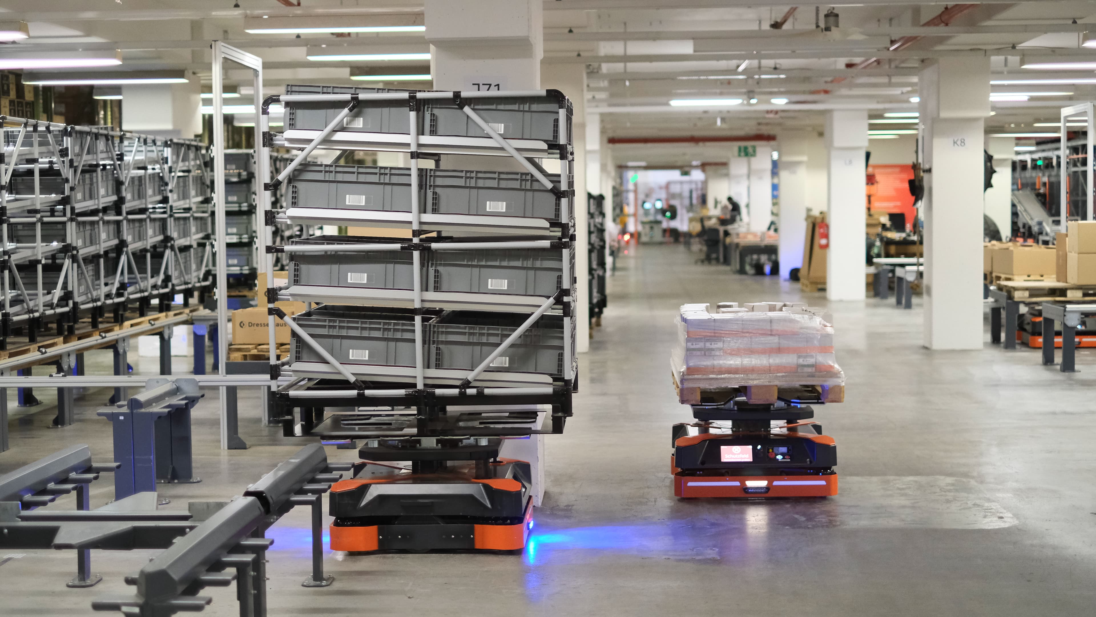 A warehouse with automated robots moving shelves and pallets of goods. The robots are orange and black, navigating the spacious, well-lit aisles. Shelves are stacked with gray bins, and some boxes are wrapped in clear plastic on pallets.