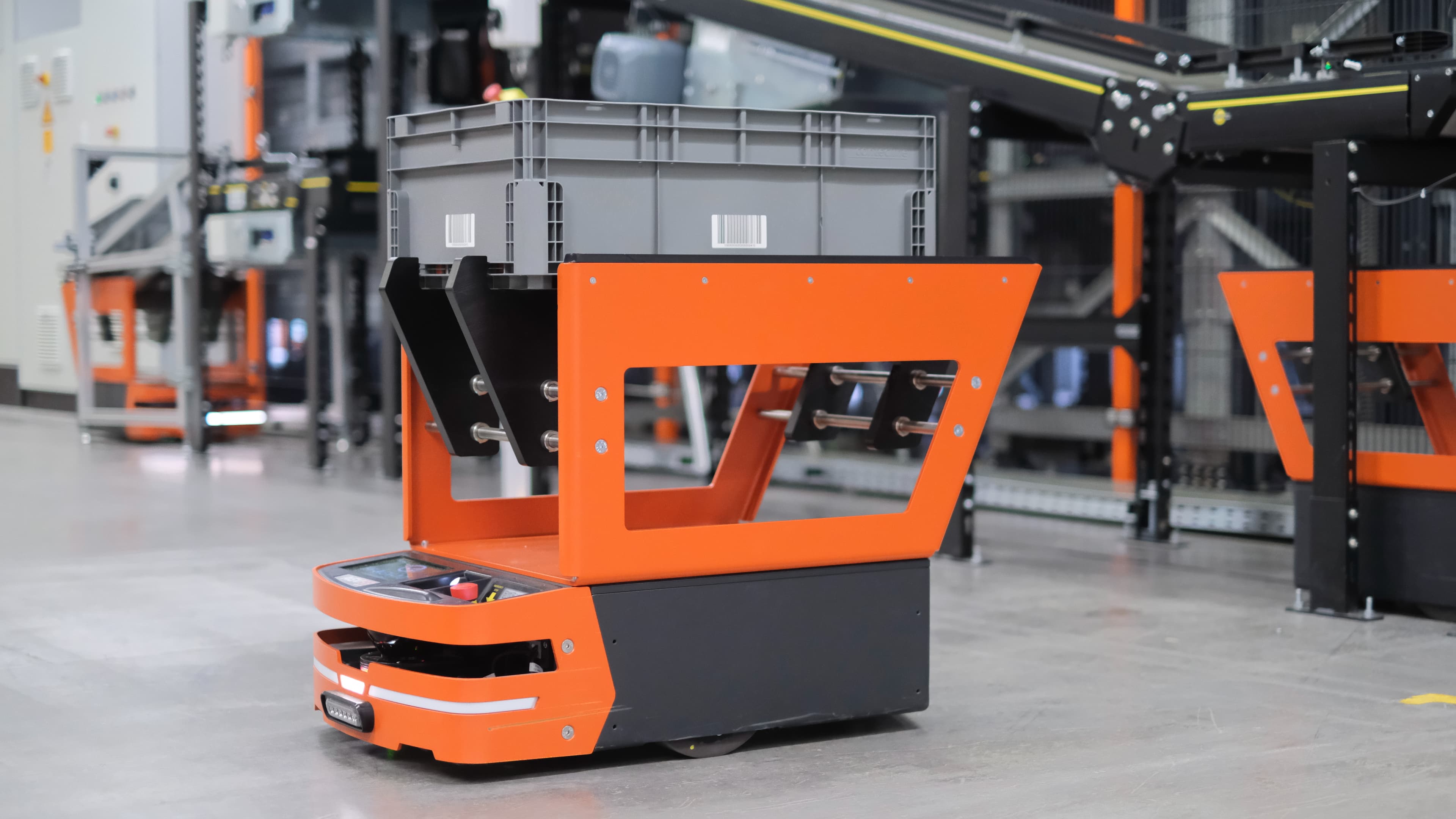 An automated guided vehicle (AGV) with an orange frame transports a gray storage container in an industrial facility. The background shows conveyor belts and machinery, emphasizing an automated logistics setting.