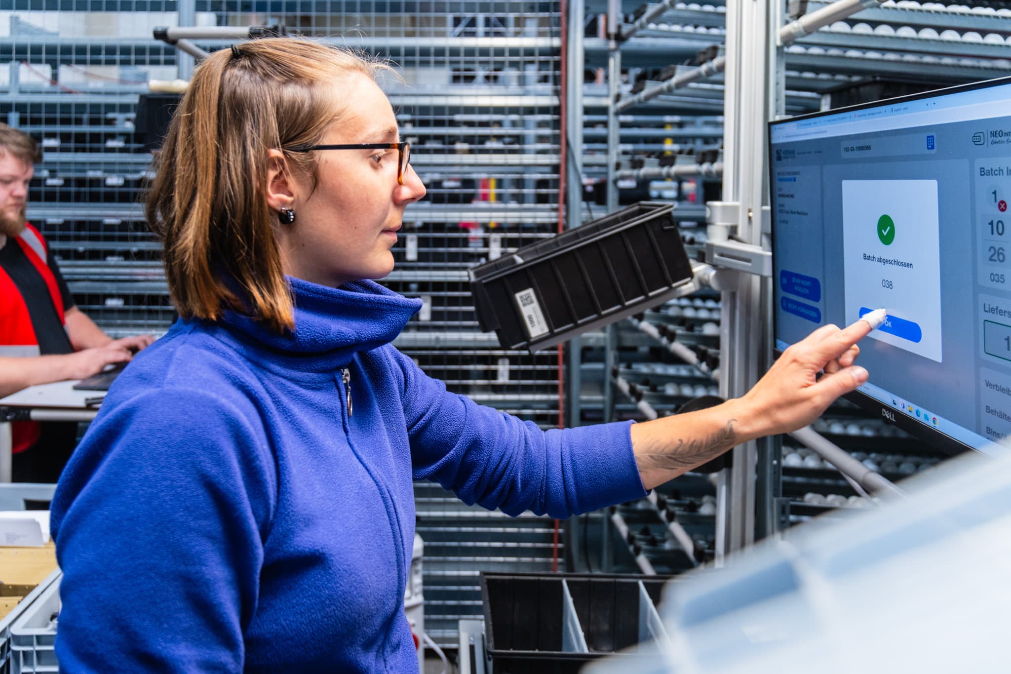 A woman in a blue sweater is using a touch screen in an industrial setting, interacting with a display that shows a confirmation screen. A man in a red shirt works at a computer in the background. The environment has metal shelving and bins.