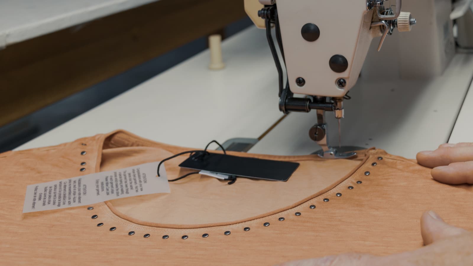A sewing machine stitching a light orange T-shirt with metal studs around the neckline. A black tag and a care label are attached to the shirt. Hands are visible guiding the fabric under the needle.