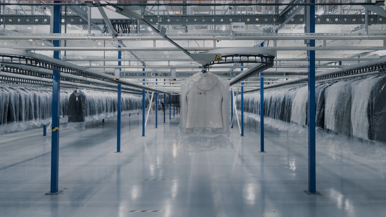Industrial warehouse interior with clothing on hangers in transparent plastic covers, suspended from a conveyor system. The space is lit with bright overhead lighting, and rows of garments extend into the background.