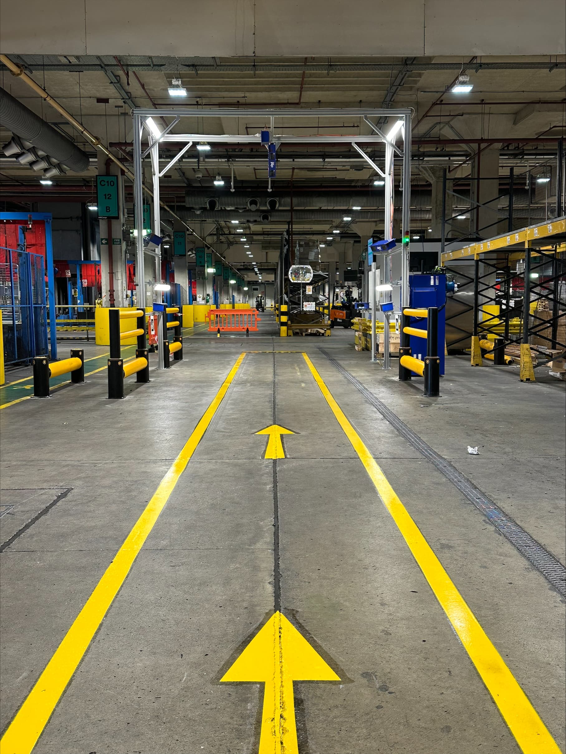 Industrial warehouse interior with a clear pathway marked by yellow lines and arrows on the concrete floor. The space is well-lit, featuring shelving units, equipment, safety barriers, and a high ceiling with exposed structures.
