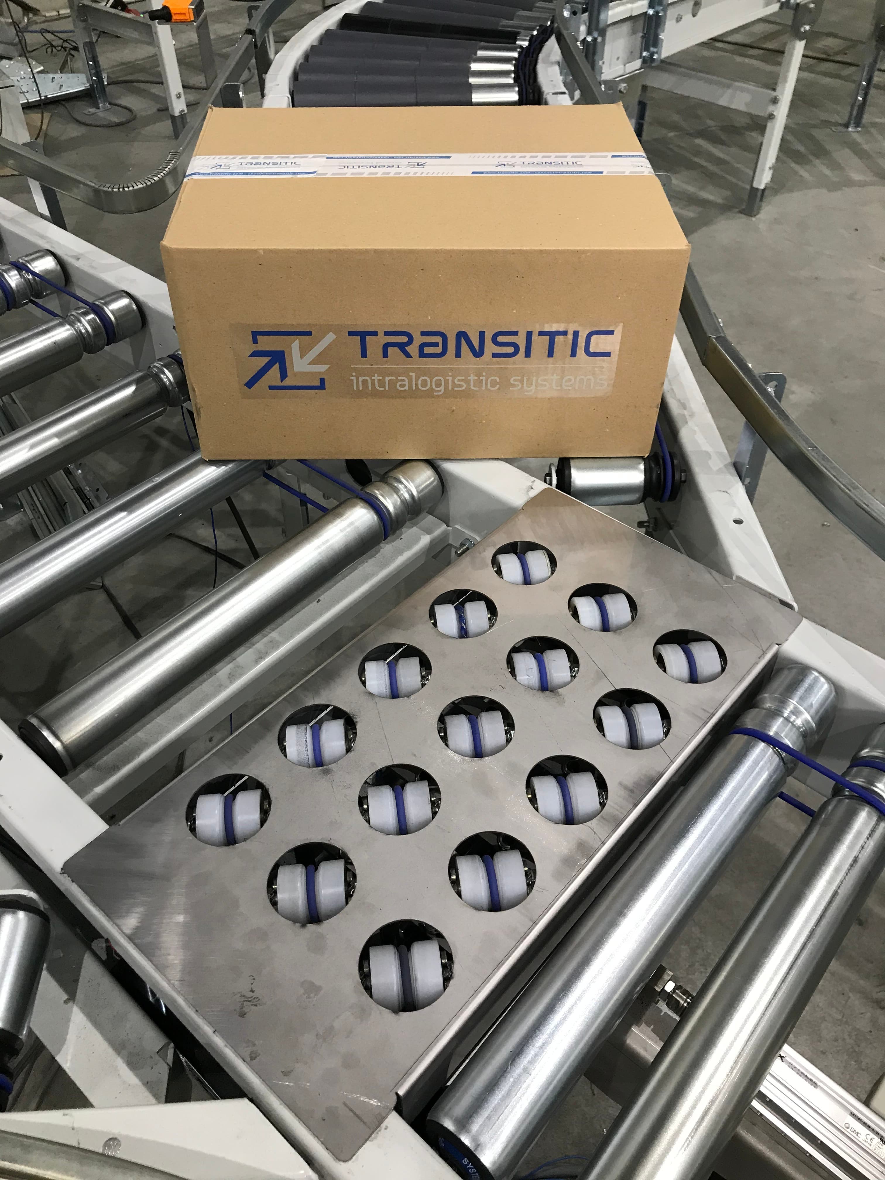 A cardboard box with TRANSITIC intralogistic systems logo rests on a conveyor system in an industrial setting. The conveyor has multiple rollers and a metal platform with adjustable wheels.