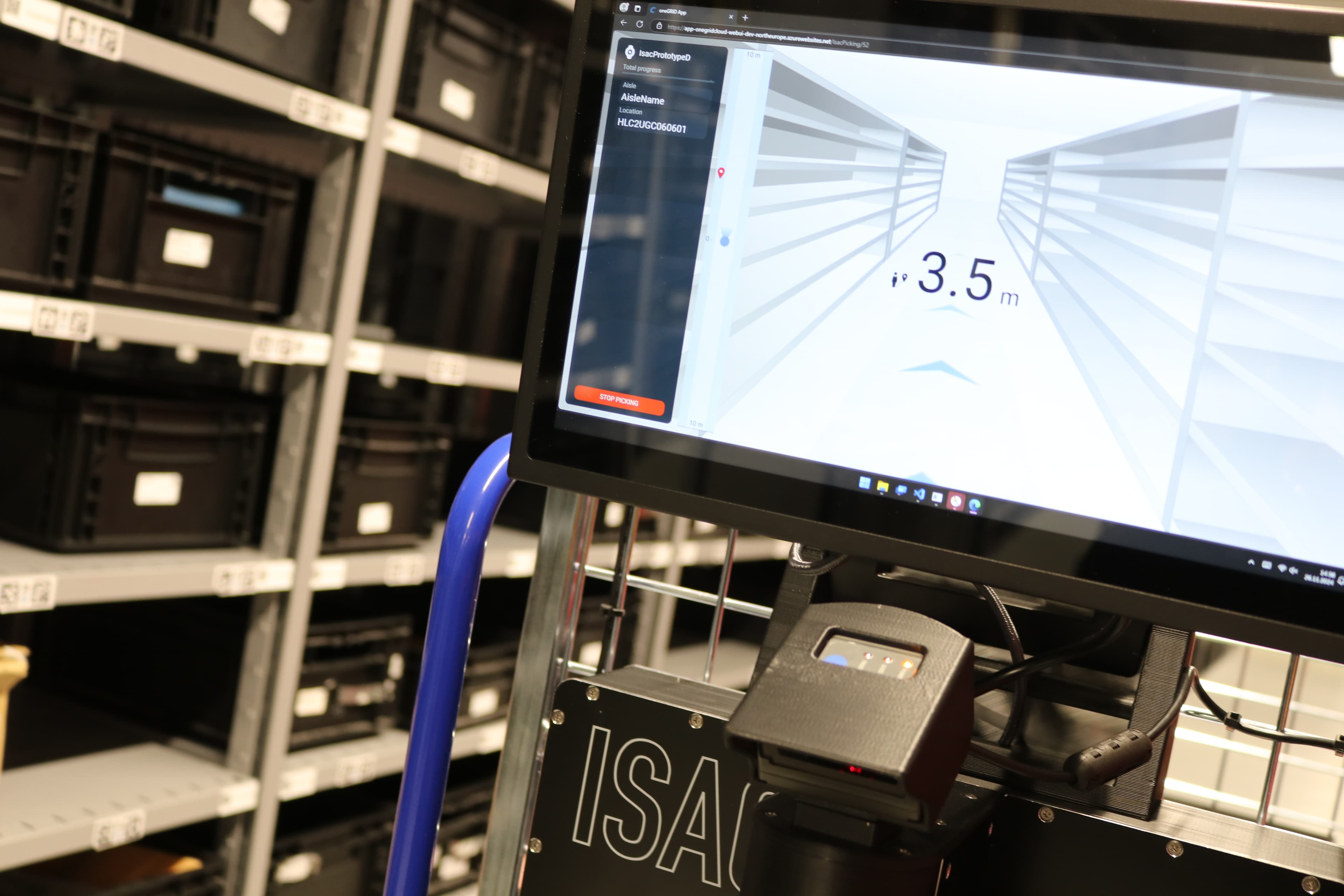 A monitor on a cart in a storage area displays a digital measurement of 3.5 m with a graphic of empty shelves. Nearby, shelves are filled with black containers labeled with white numbers.