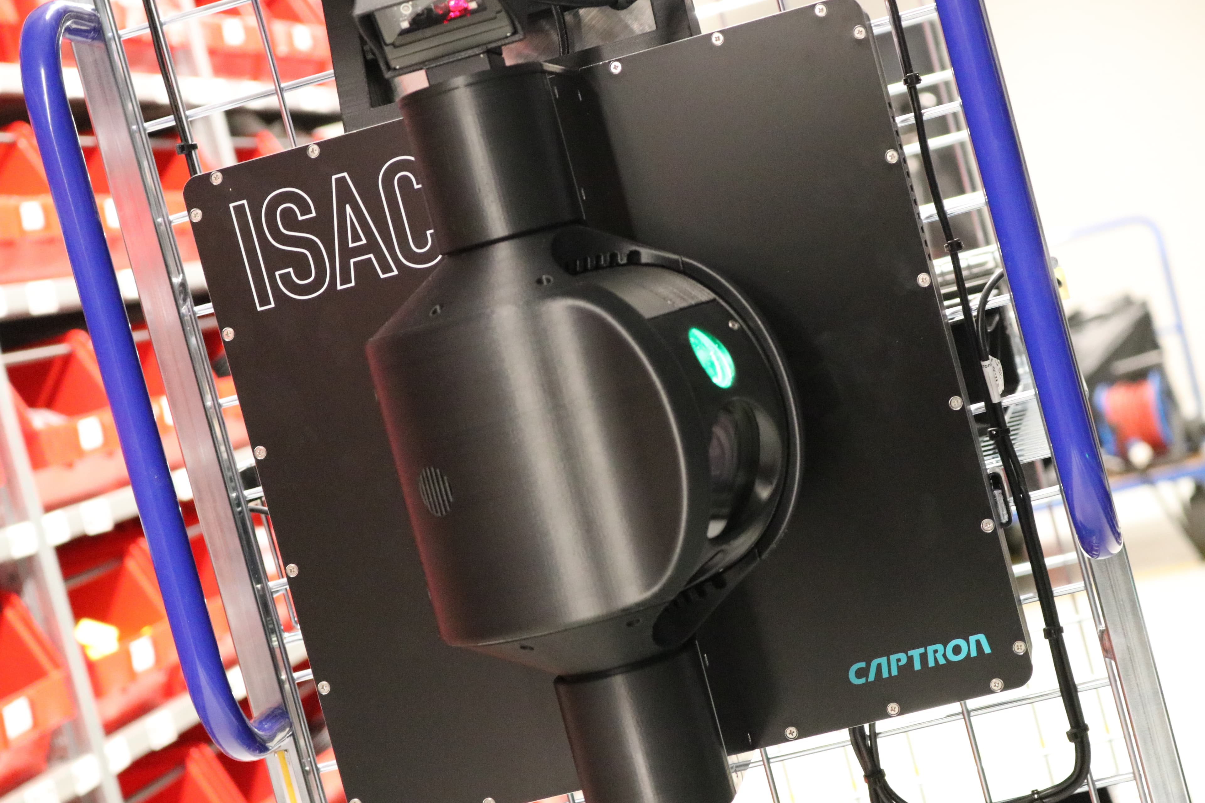 Close-up of a black device with ISAC and CAPTRON labels mounted on a metal frame, surrounded by red bins. The device features a cylindrical component and a glowing green button, suggesting industrial or technological use.
