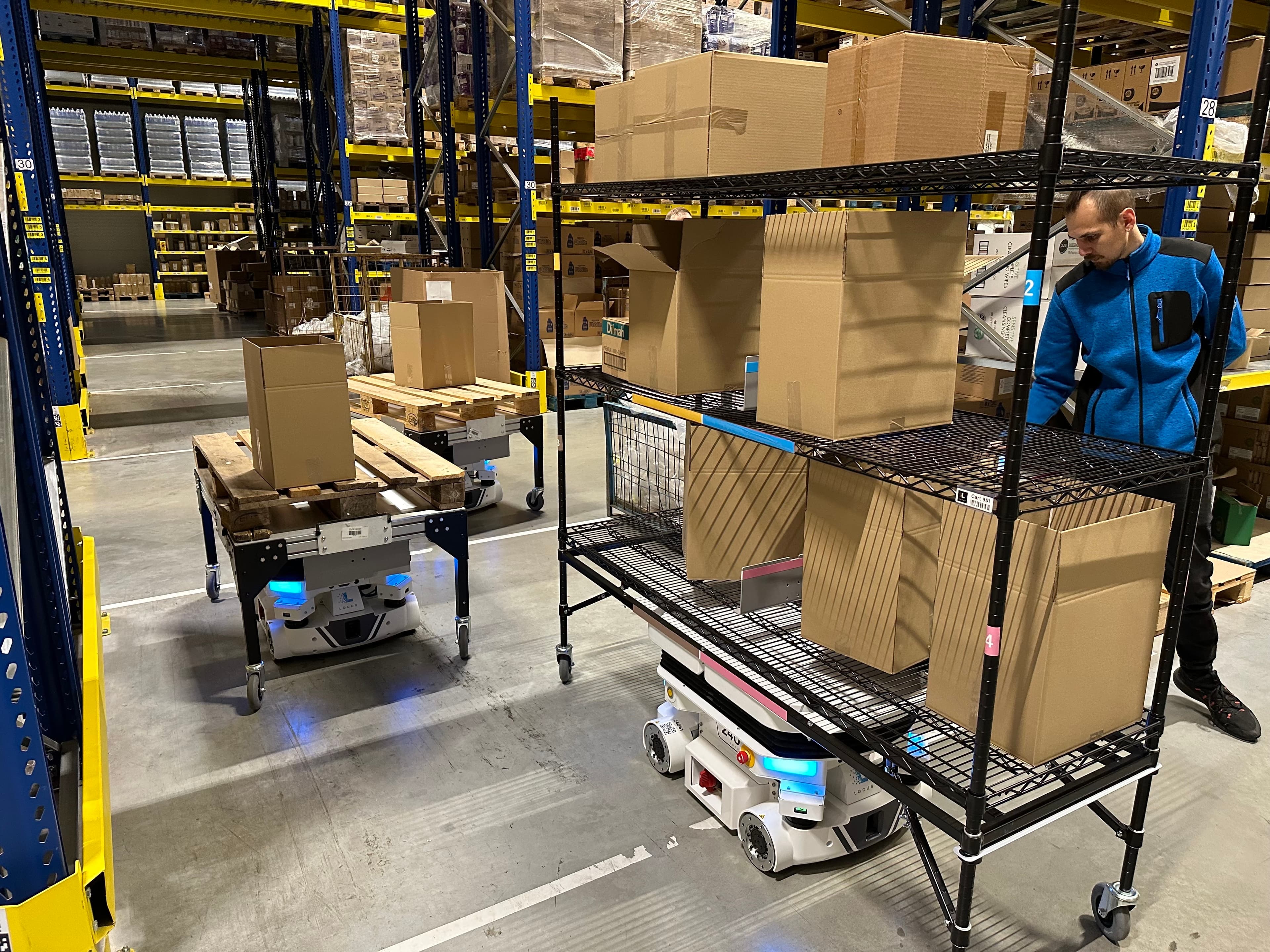 A man in a blue jacket is in a warehouse, organizing boxes on a metal shelf. Two robotic carts, carrying parcels, are moving along the aisle. Shelves are stocked with various packages and are stacked high in the background.