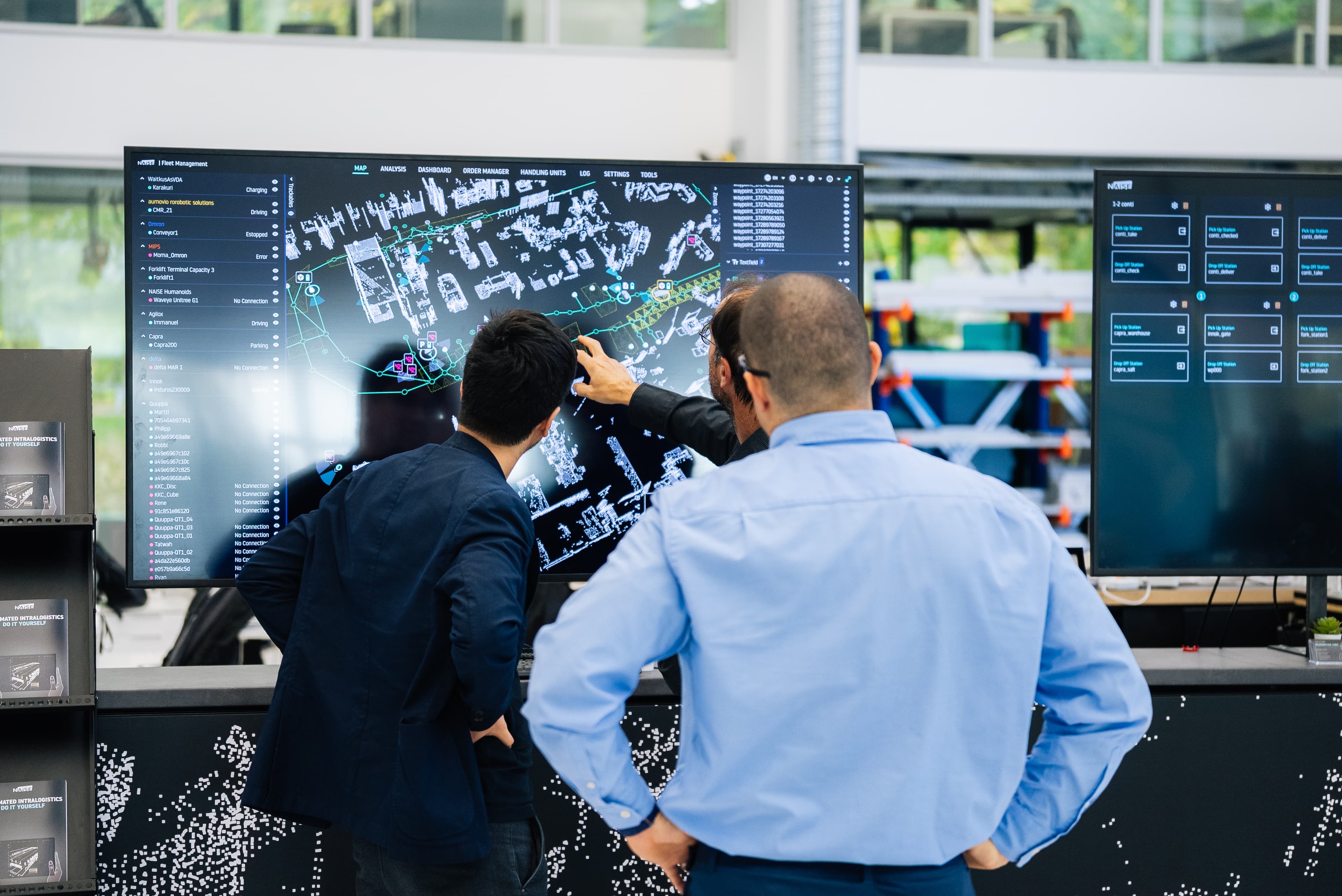 Drei Personen in einem modernen Büro sehen sich eine große digitale Logistikkarte auf einem Bildschirm an und arbeiten gemeinsam an der Optimierung von Sendungen und Routen.