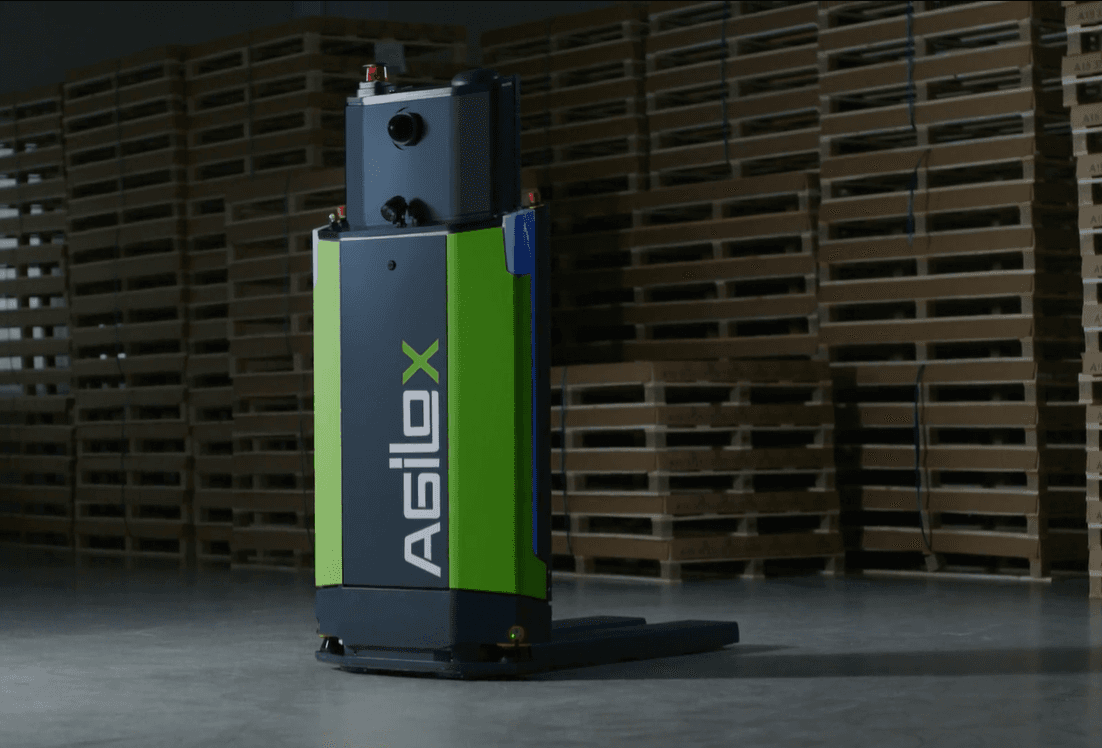 Ein grün-schwarzer autonomer Agilox-Roboter navigiert durch die Lagerhalle, im Hintergrund stehen gestapelte Holzpaletten für den Logistikbetrieb bereit.