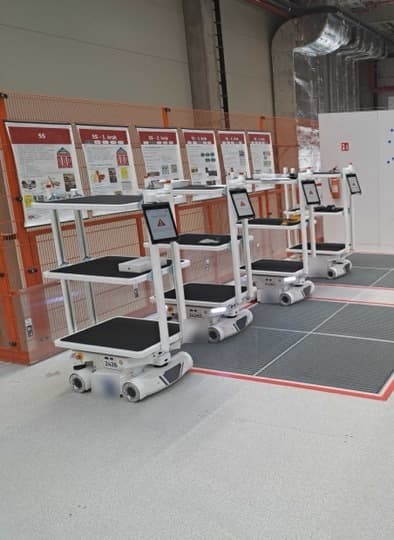 Robotic carts with shelves lined up in a warehouse setting, each equipped with a touchscreen interface. They are positioned near informational posters on a metal grid floor, surrounded by safety barriers and industrial equipment.