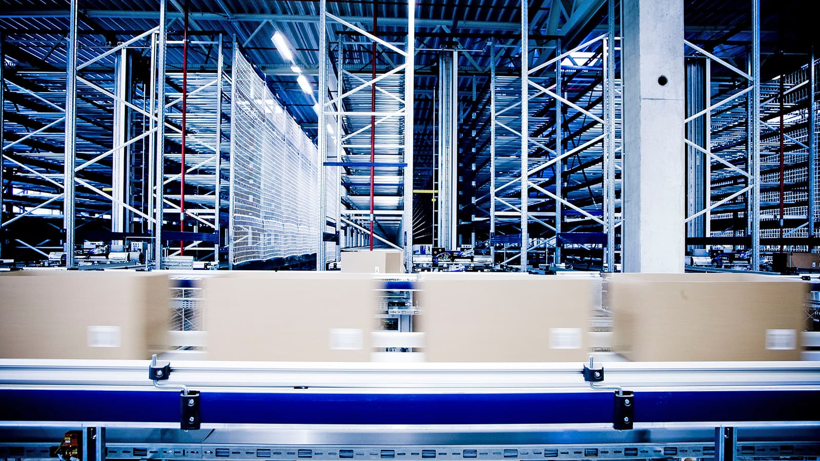 Blurred moving boxes on a conveyor belt in a large warehouse with metal shelving and industrial lighting.