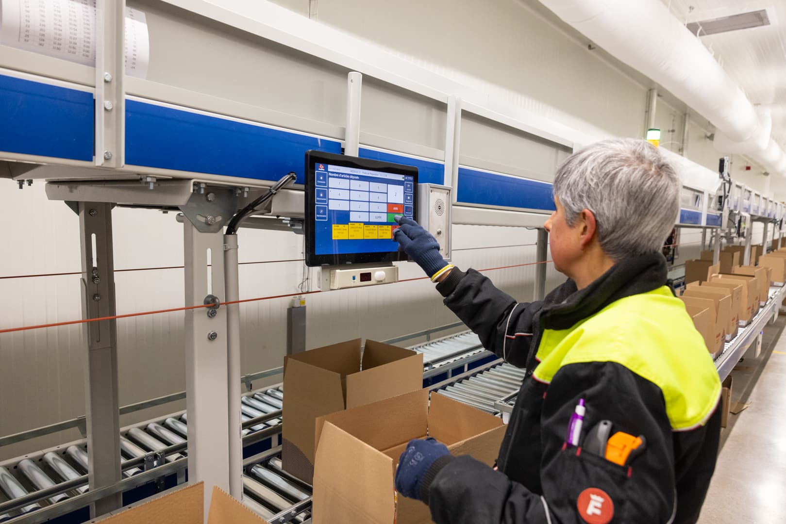 A worker wearing a high-visibility jacket operates a touchscreen control panel on a conveyor belt system in a warehouse. Cardboard boxes are placed on the conveyor, suggesting a packaging or distribution process.