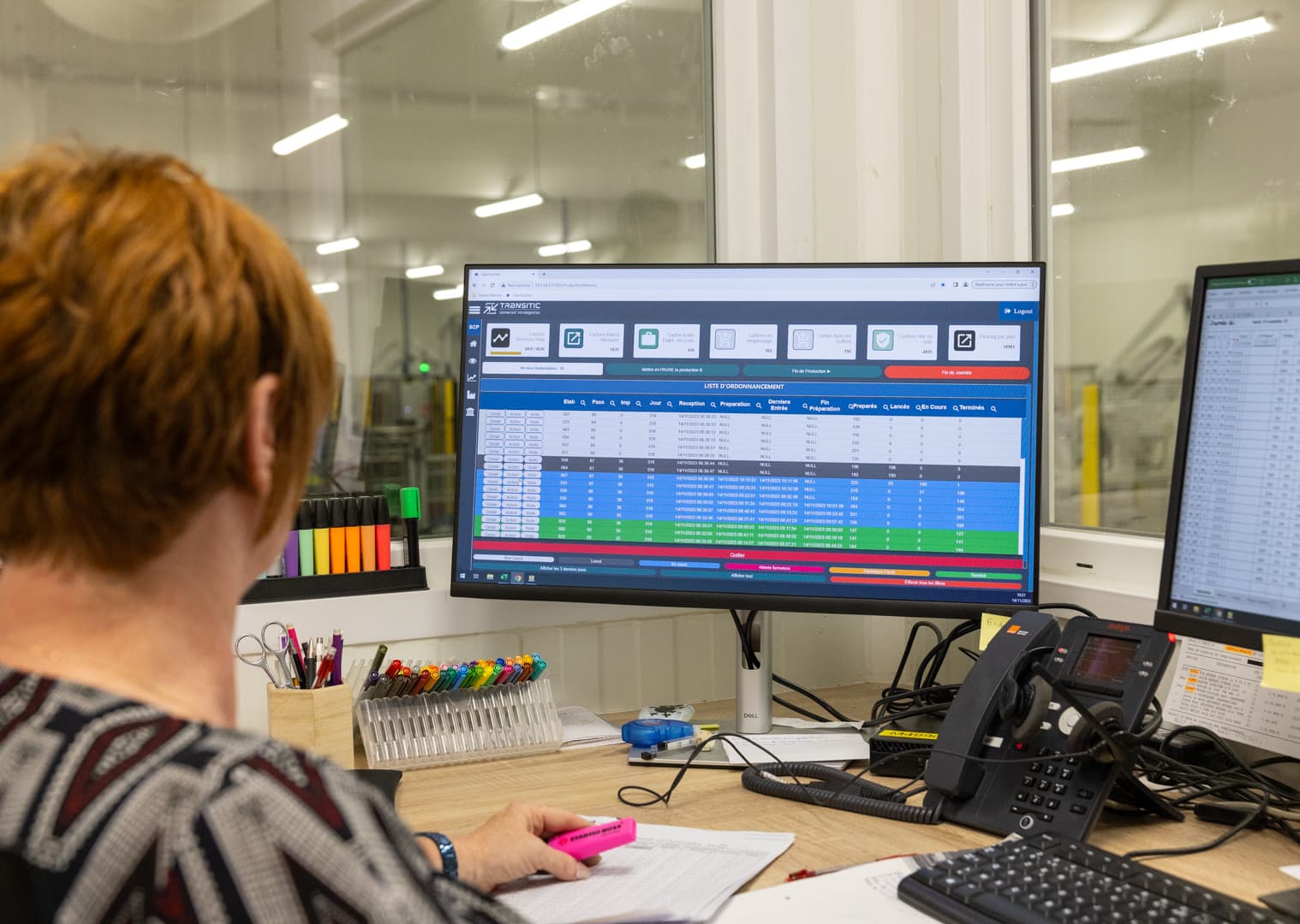 A person with short red hair sits at a desk working on a computer. The screen displays a colorful spreadsheet. Office supplies, a phone, and papers clutter the desk. Large windows in the background show an industrial workspace.