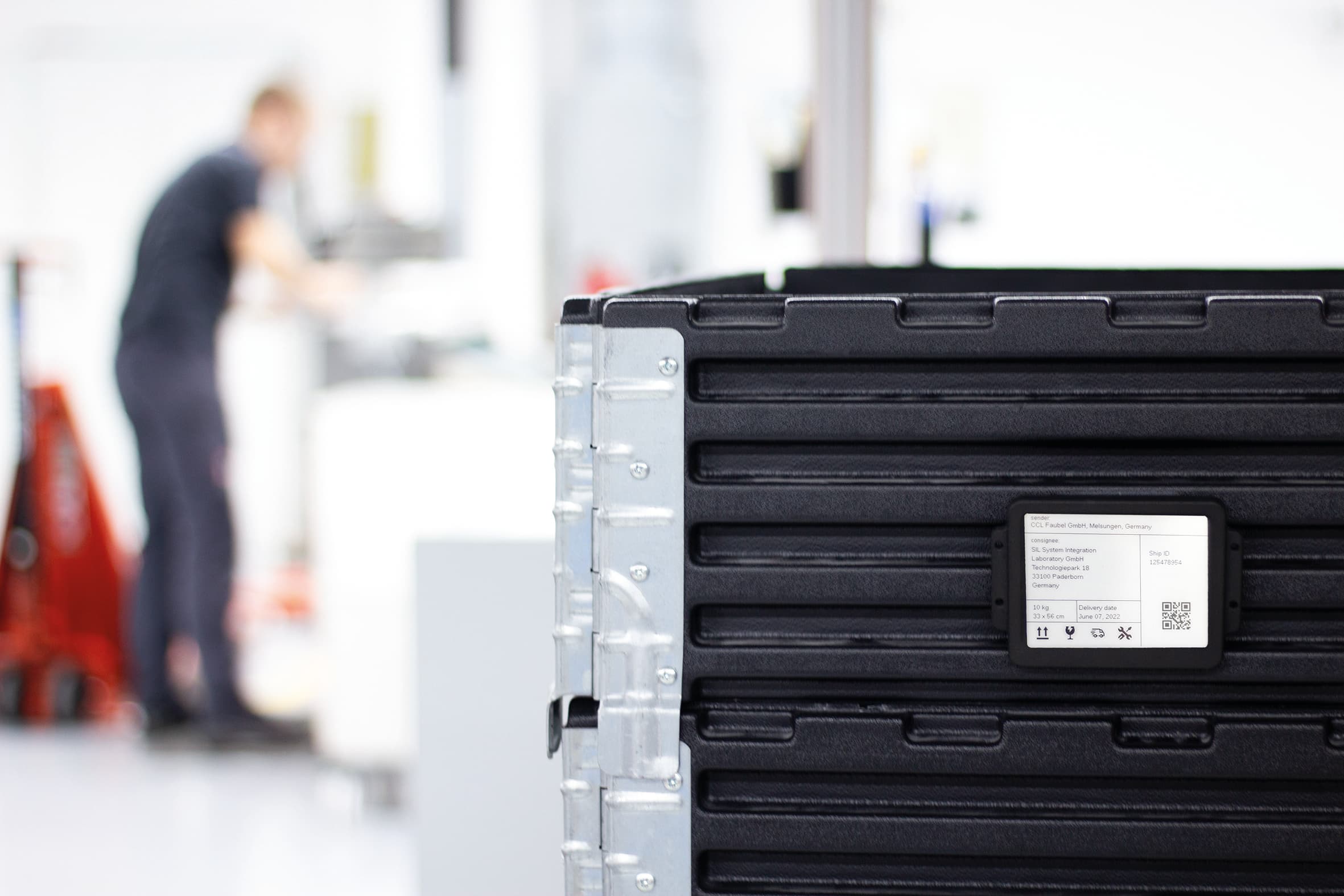 Schwarze Transportkiste mit Versandetikett von CCL Faubel GmbH im Vordergrund, Mitarbeiter in Produktionshalle unscharf im Hintergrund