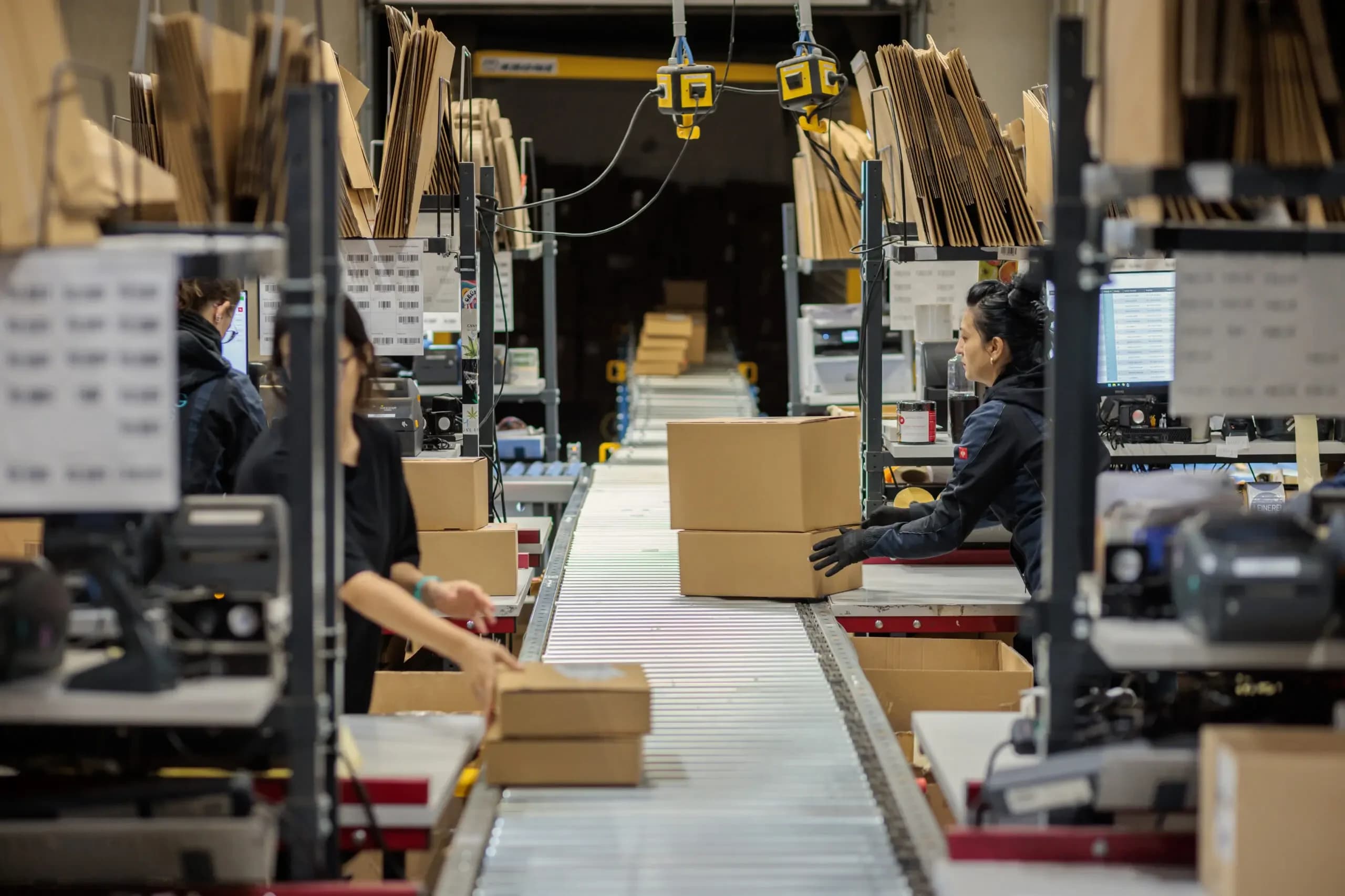 Mitarbeiterinnen an Förderband-Packstation mit Kartons und Monitoren in einem Fulfillment-Lager